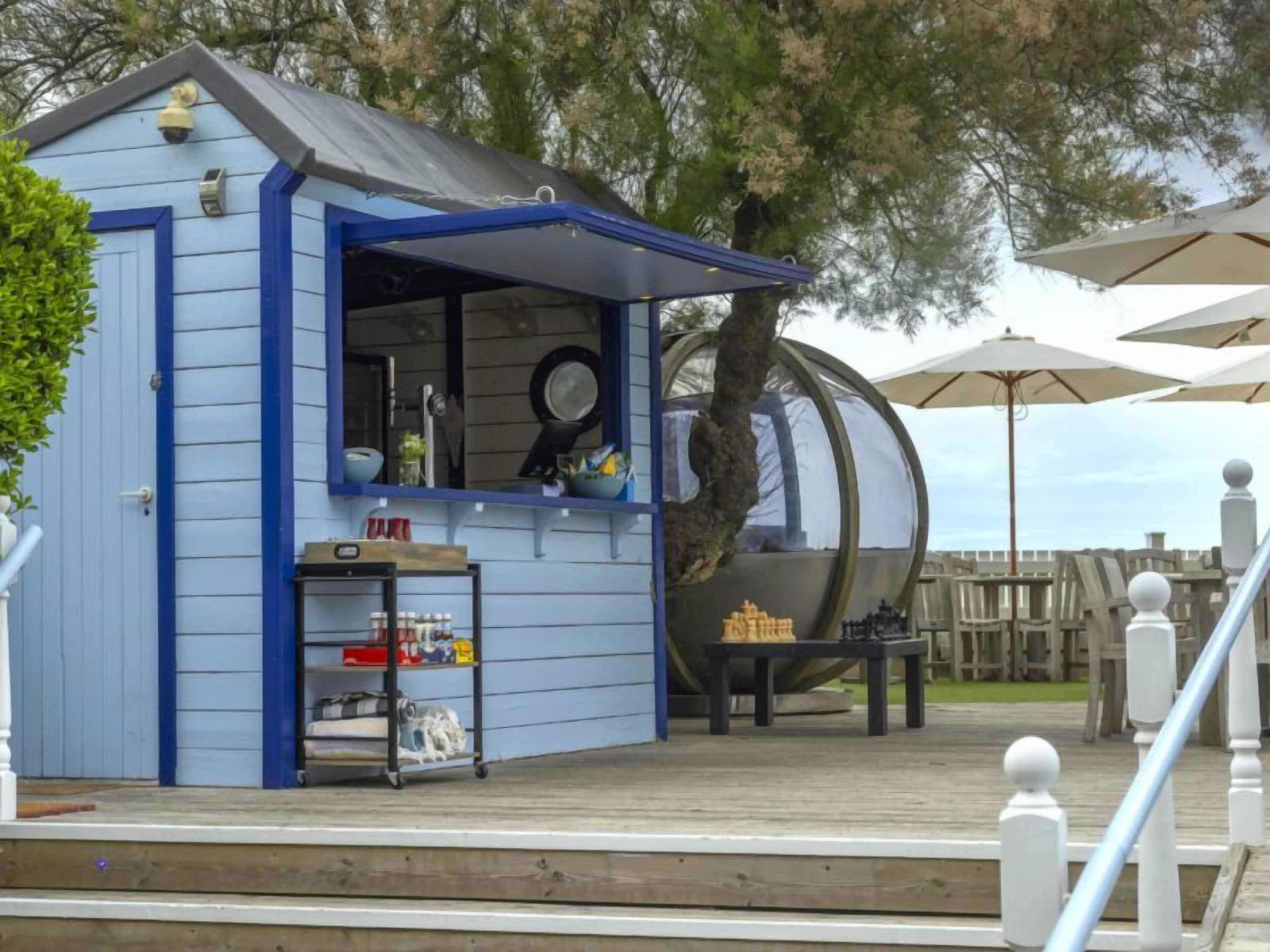 A small blue building with a glass enclosure, a wooden deck, and outdoor umbrellas.