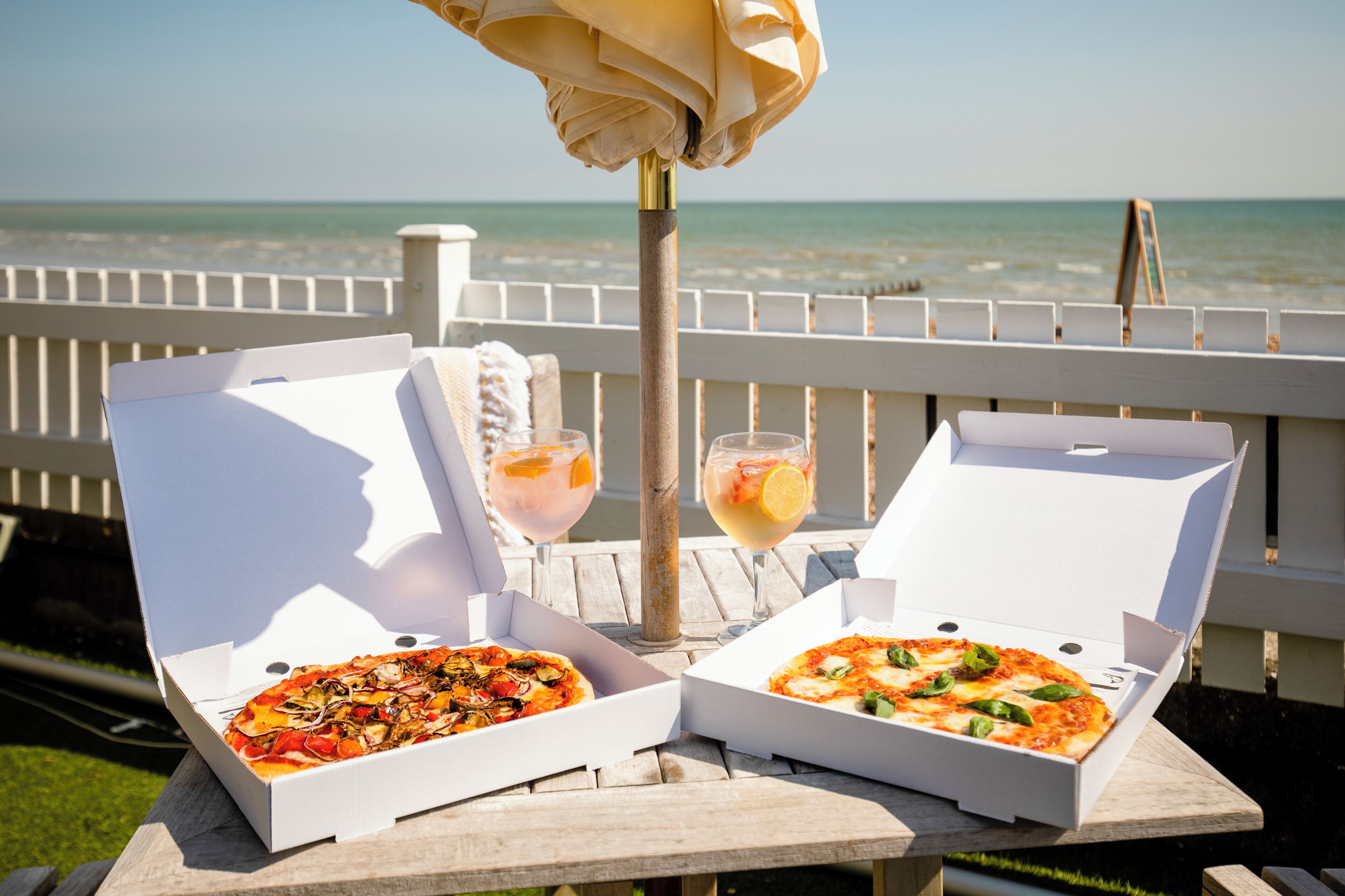 Zwei offene Pizzakartons auf einem Holztisch, im Hintergrund Blick auf Strand und Meer.