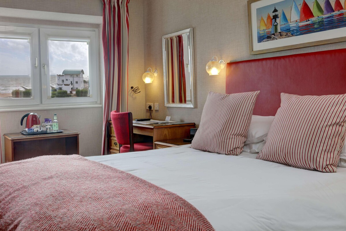 A hotel room with a large bed, a desk, a chair, and a view of the sea through a window.