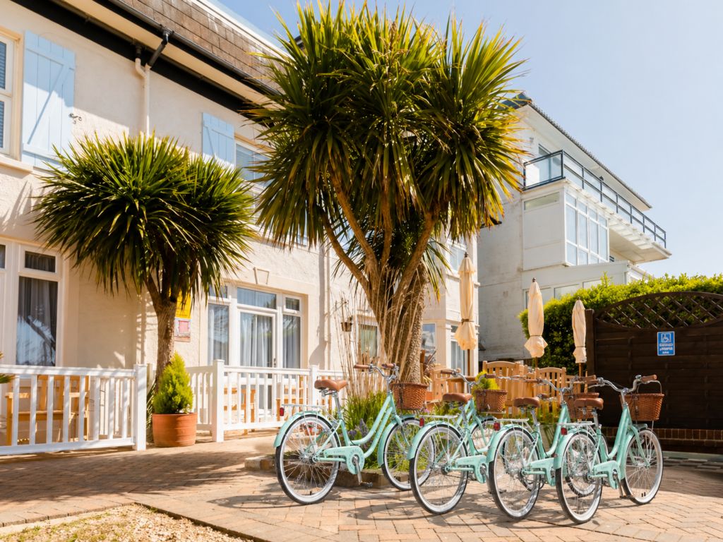 Une rangée de vélos est garée devant un immeuble à la façade blanche et aux volets bleus.