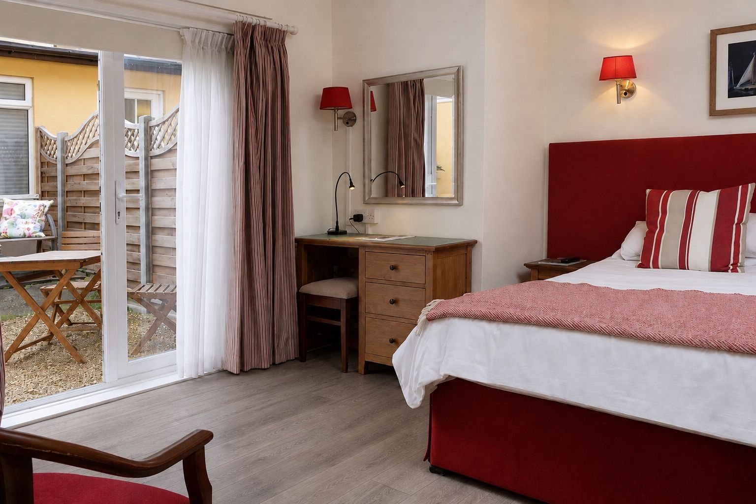 A bedroom with a bed, a desk, a chair, a mirror, and a sliding glass door leading to an outdoor area.