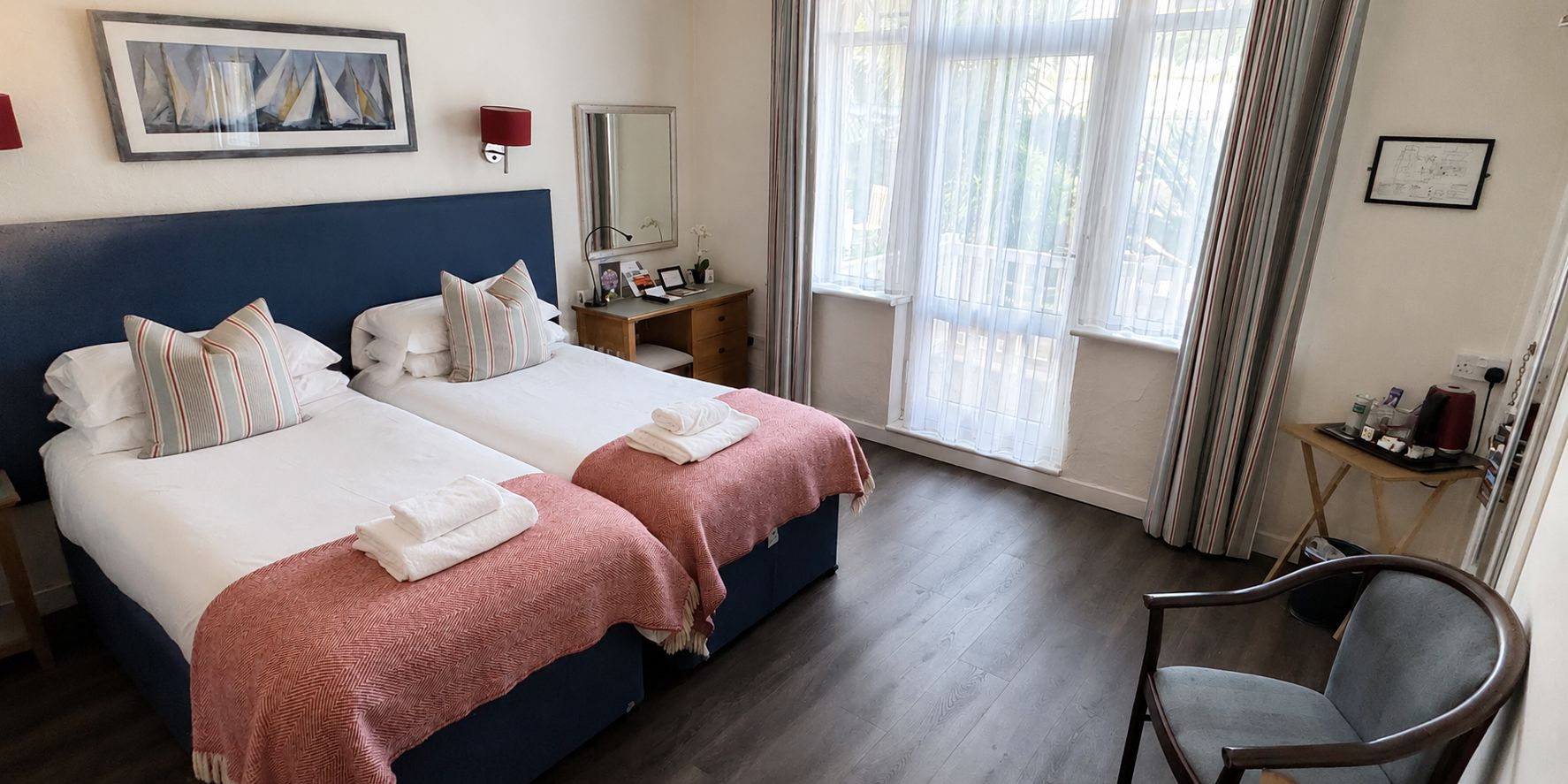 A hotel room with two beds, a desk, a chair, a window with curtains, and a painting on the wall.