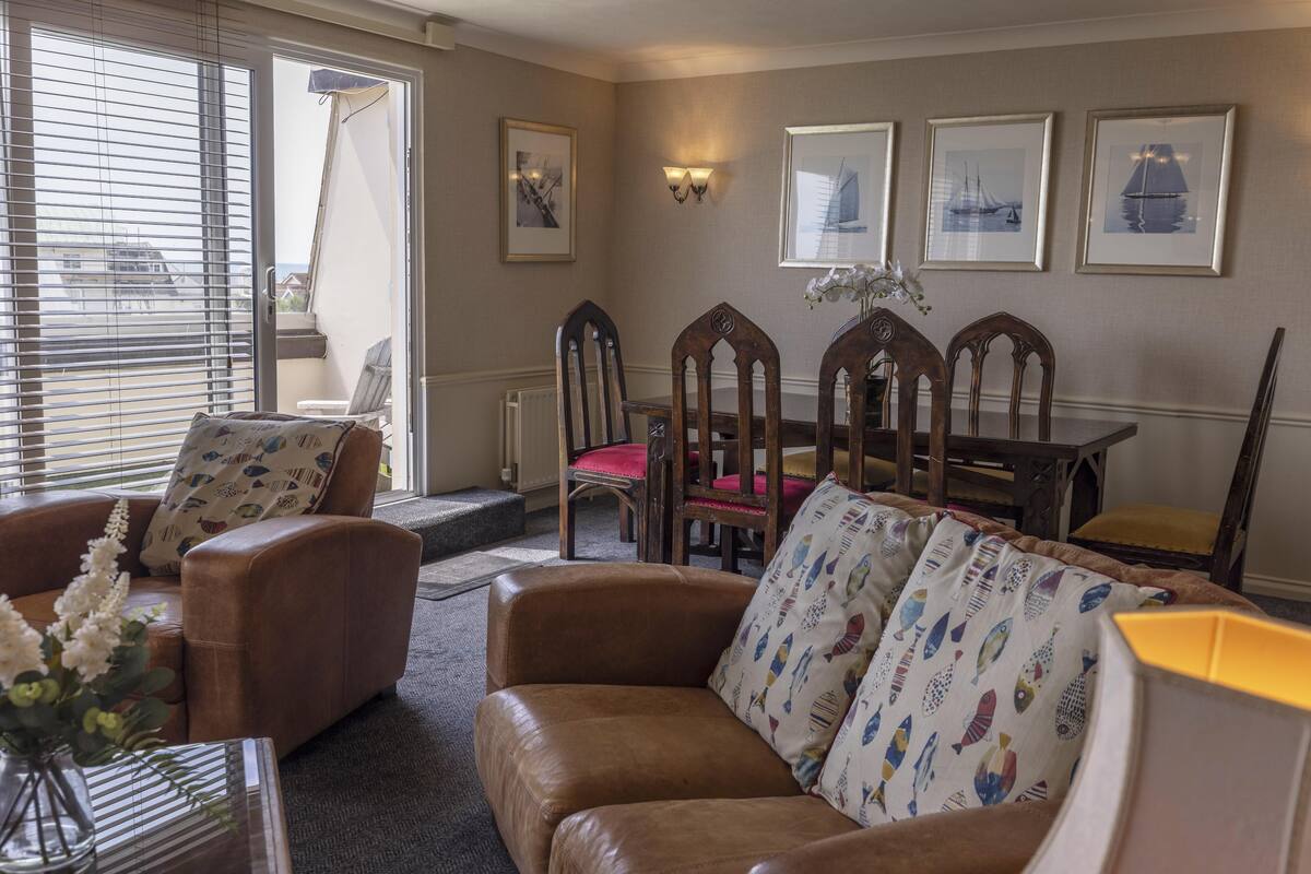 A living room with a brown leather sofa, a dining table with chairs, and a view through a sliding door to the outside.