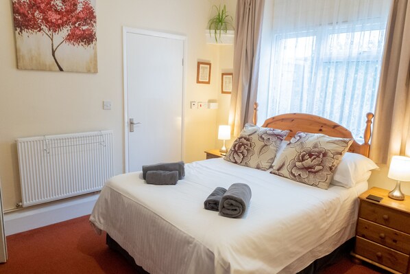 A neatly made bed with a wooden headboard, two bedside lamps, and a window with curtains. - Beecroft Lodge (Paignton)