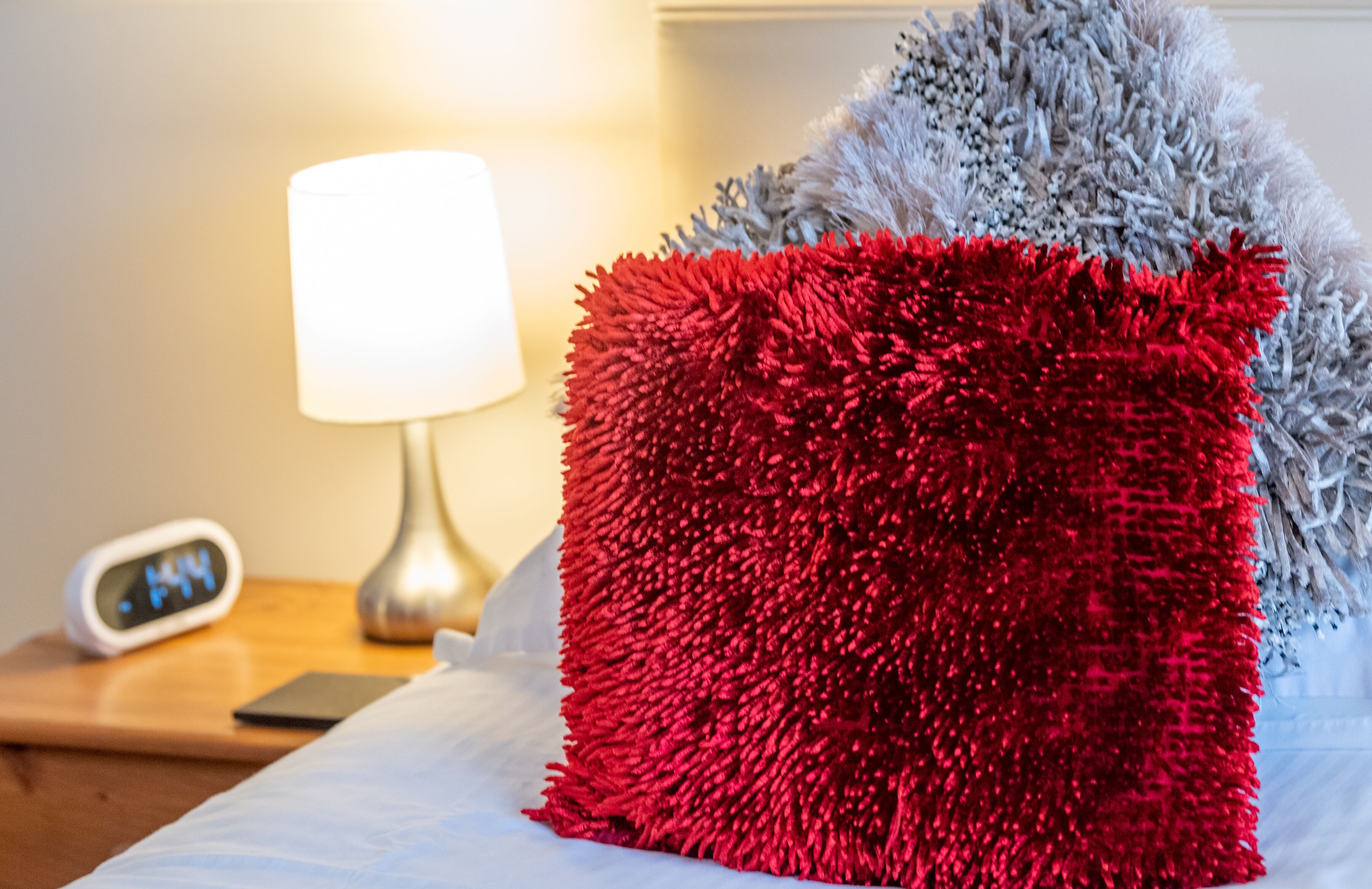 A bed with a red and grey textured pillow, a bedside lamp, and a digital clock on a wooden nightstand.