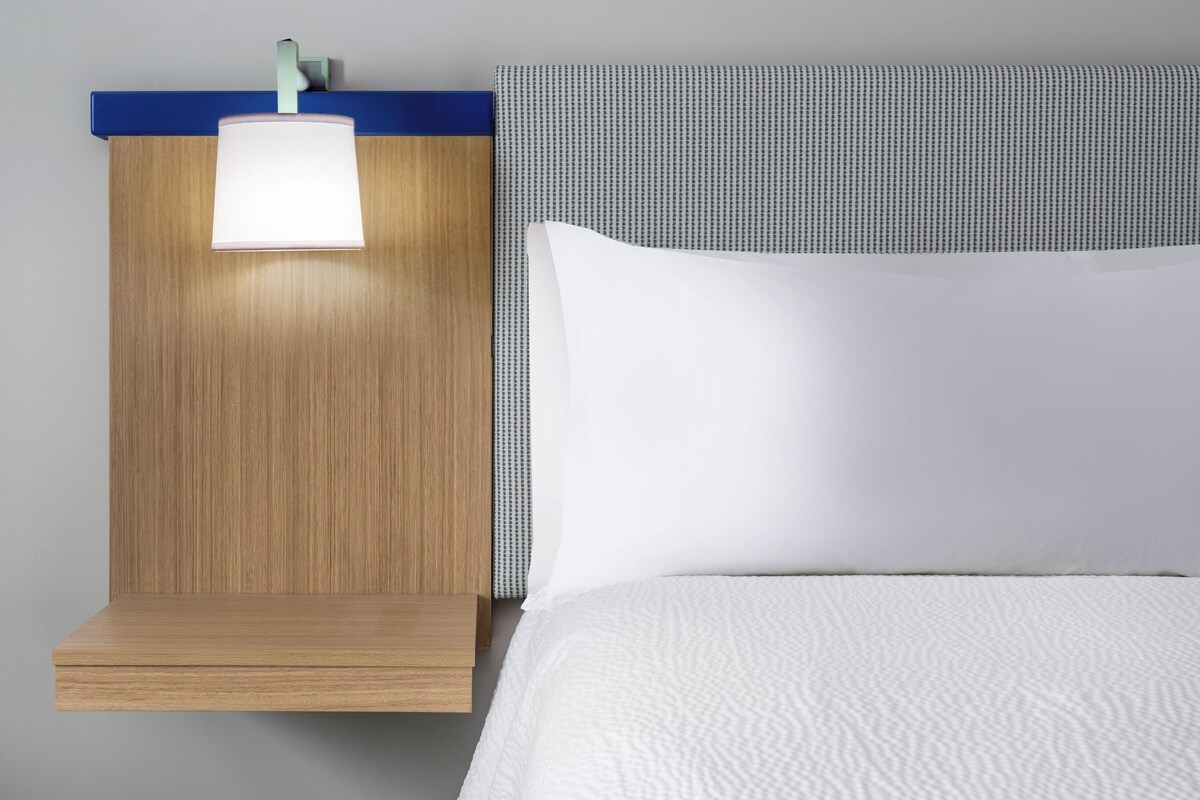 Ein Hotelzimmer mit einem Holznachttisch, einer weißen Lampe, einem weißen Bett und einem blauen Kopfteil.