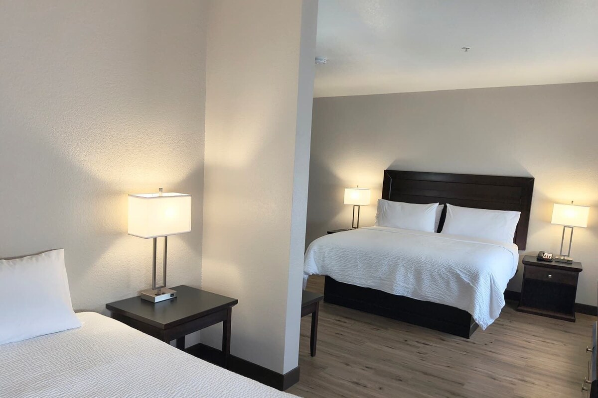 Una habitación de hotel con una cama, dos mesitas de noche con lámparas, suelo de madera y una pared lisa.