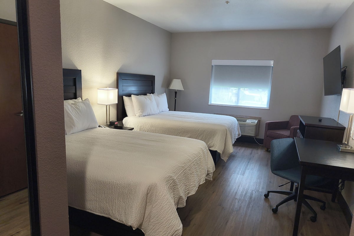 Quarto de hotel com duas camas, uma escrivaninha, uma cadeira, uma janela e televisão na parede.