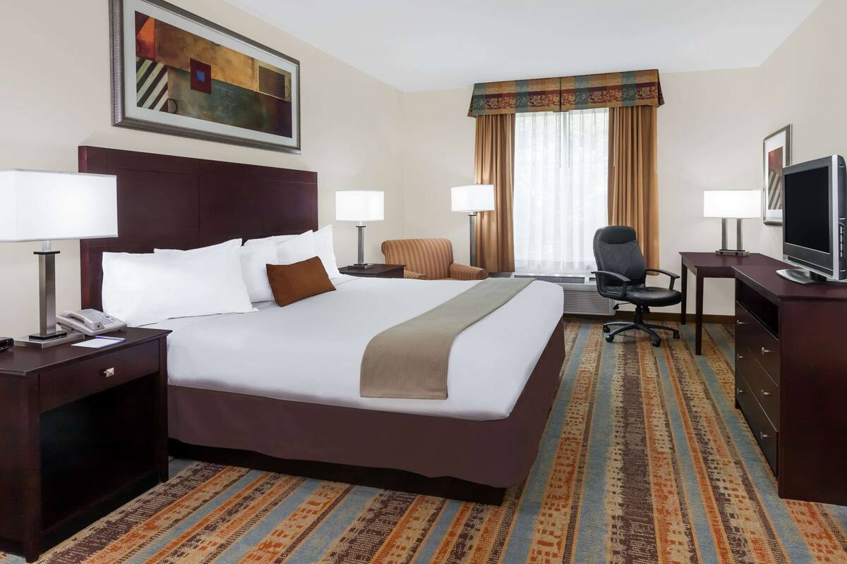 A hotel room with a large bed, two bedside tables with lamps, a desk with a chair, a television, and a window with curtains.