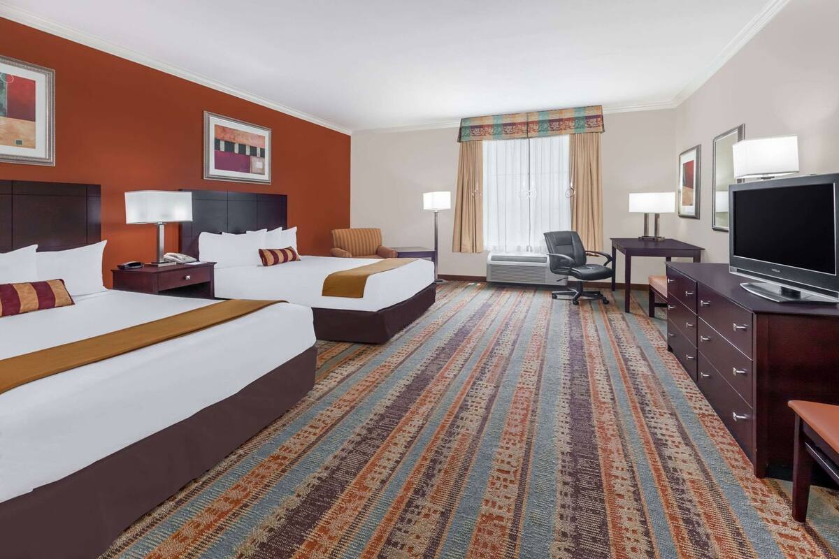 A hotel room with two beds, a desk, a television, and a large window with curtains.