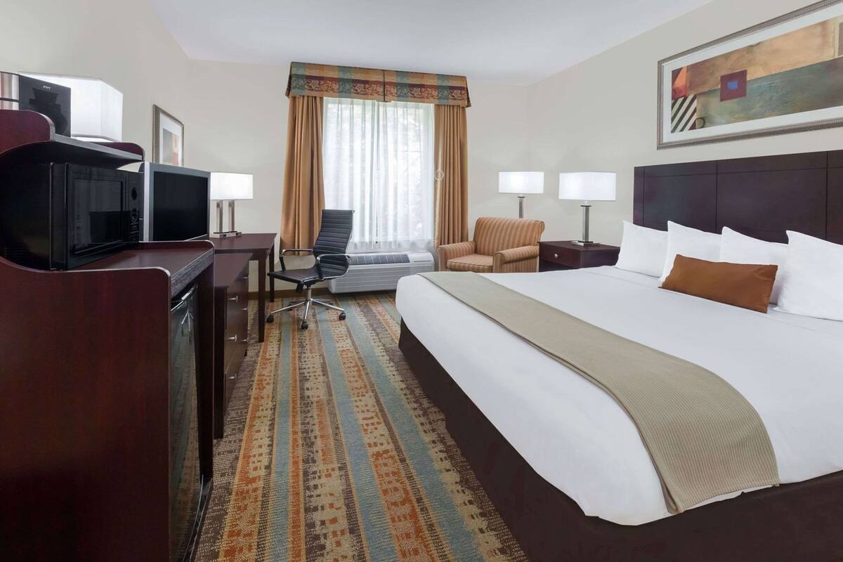 A hotel room with a bed, two chairs, a desk, a TV, and a window with curtains.