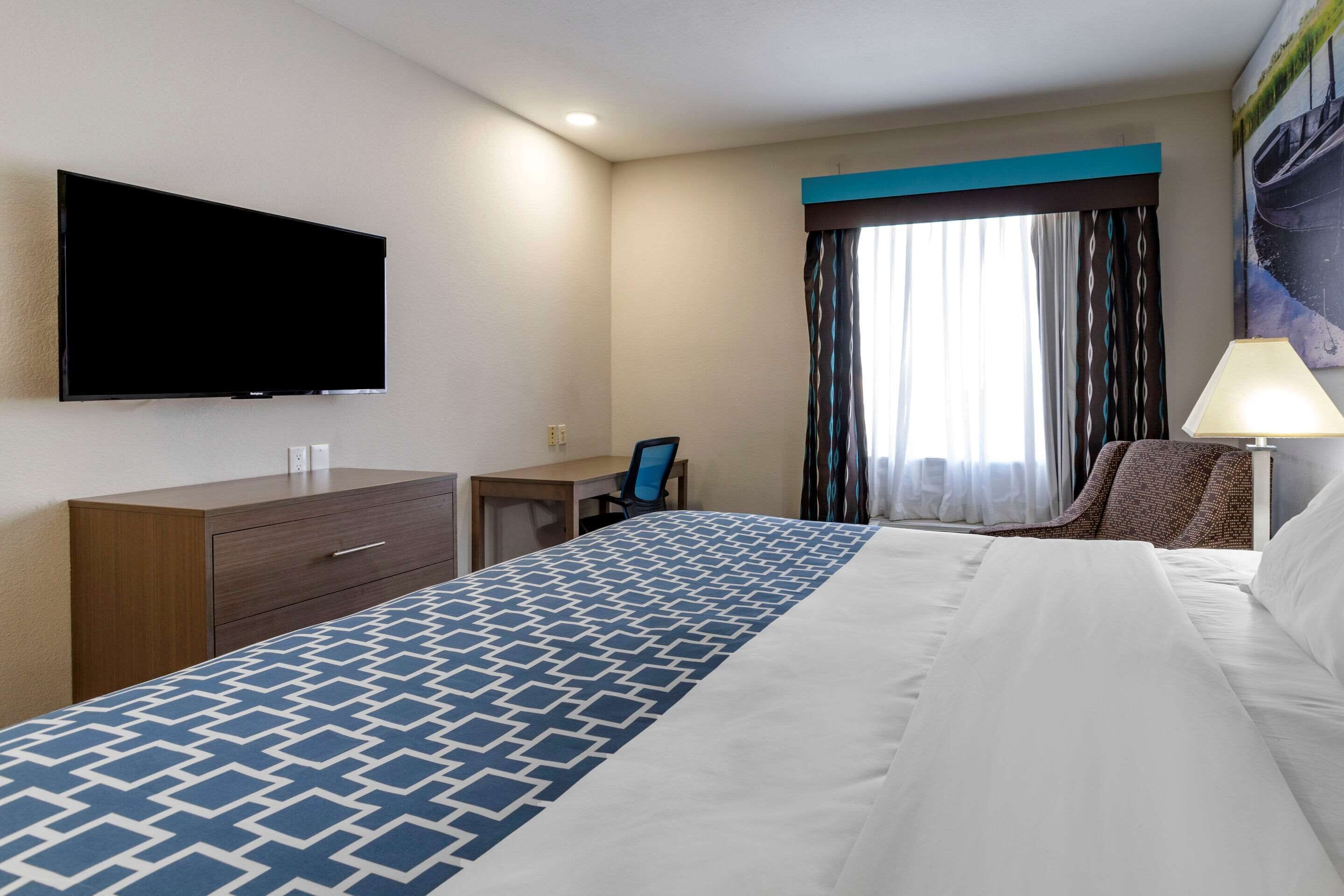 A hotel room with a large bed, a flat-screen TV mounted on the wall, a desk with a chair, and a window with curtains.