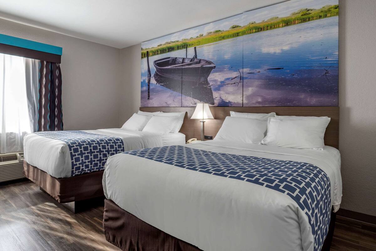 A hotel room with two beds, a large painting of a boat on water, a window with curtains, and a bedside lamp.
