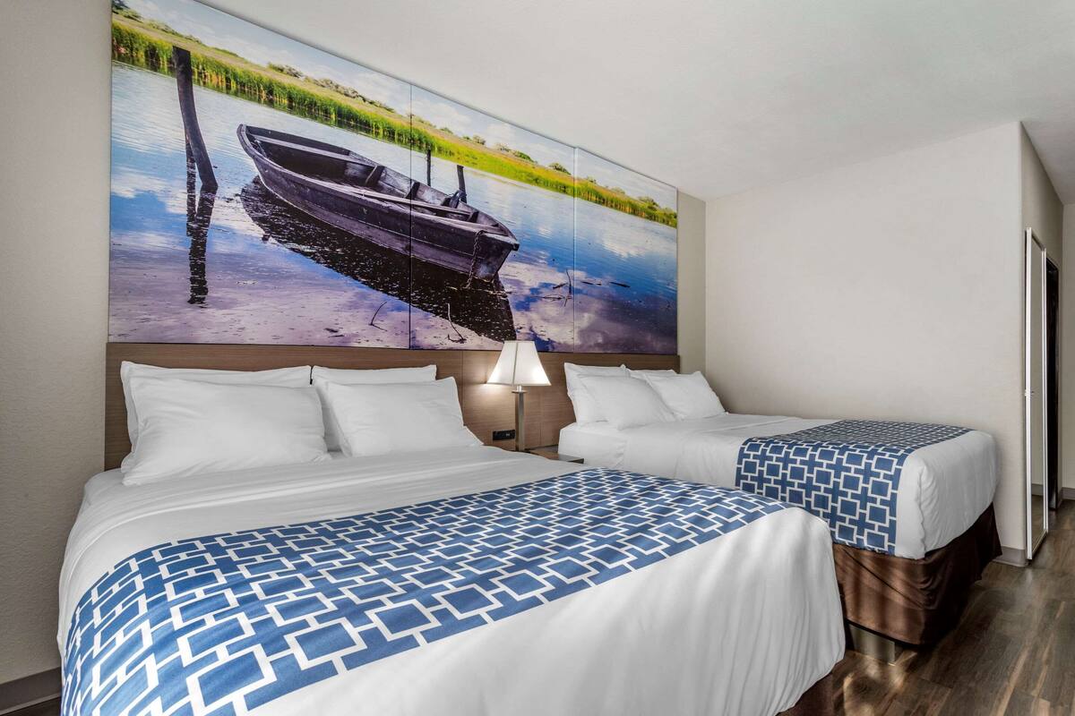 A hotel room with a large bed, two smaller beds, a painting of a boat on water, and a lamp.
