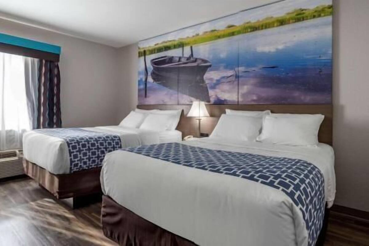 A hotel room with two beds, a painting of a boat on water, a window with curtains, and a lamp.