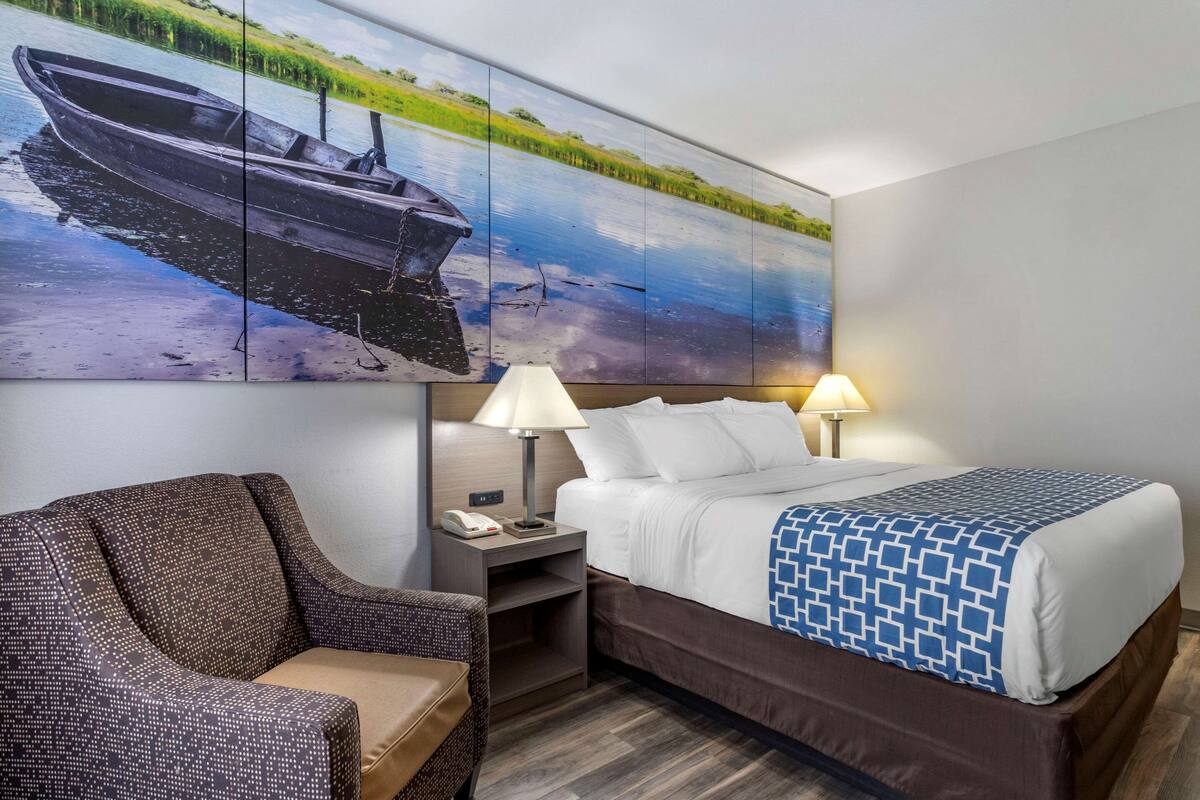 A hotel room with a bed, two bedside lamps, a nightstand, a chair, and a large landscape painting on the wall.