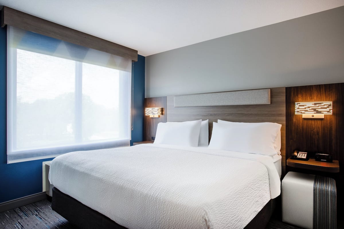 Hotellihuone, jossa on suuri sänky, yöpöytä lampun kanssa ja ikkuna, jossa on sälekaihtimet.
