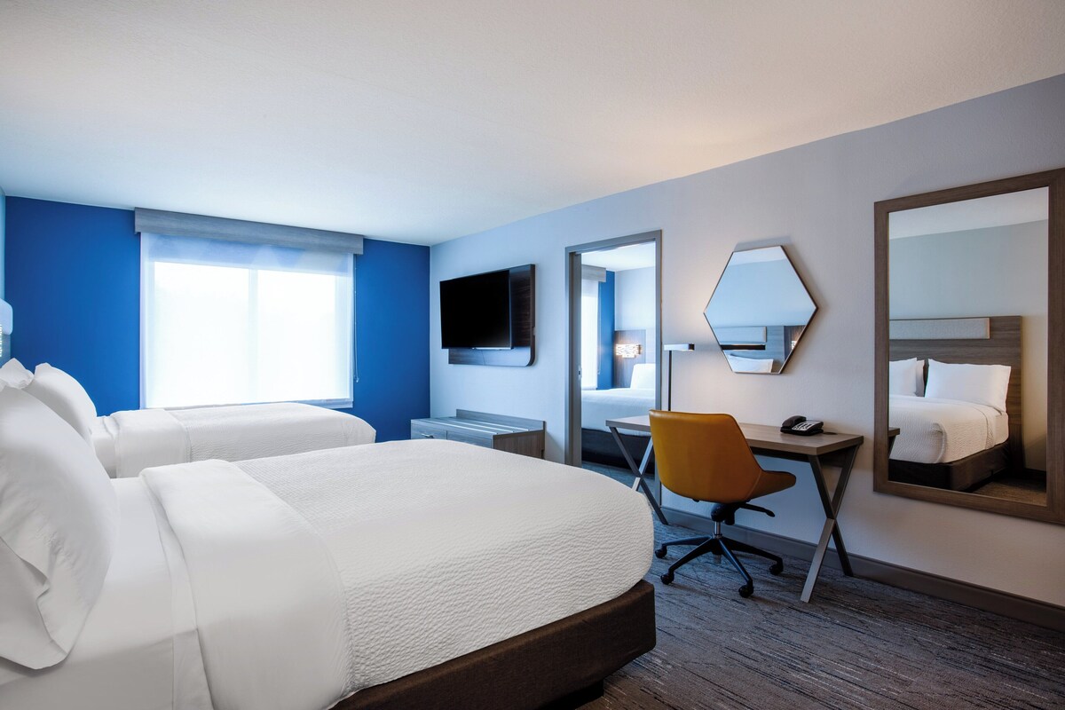 Habitación de hotel con dos camas, un escritorio, una silla, un televisor y un espejo grande.