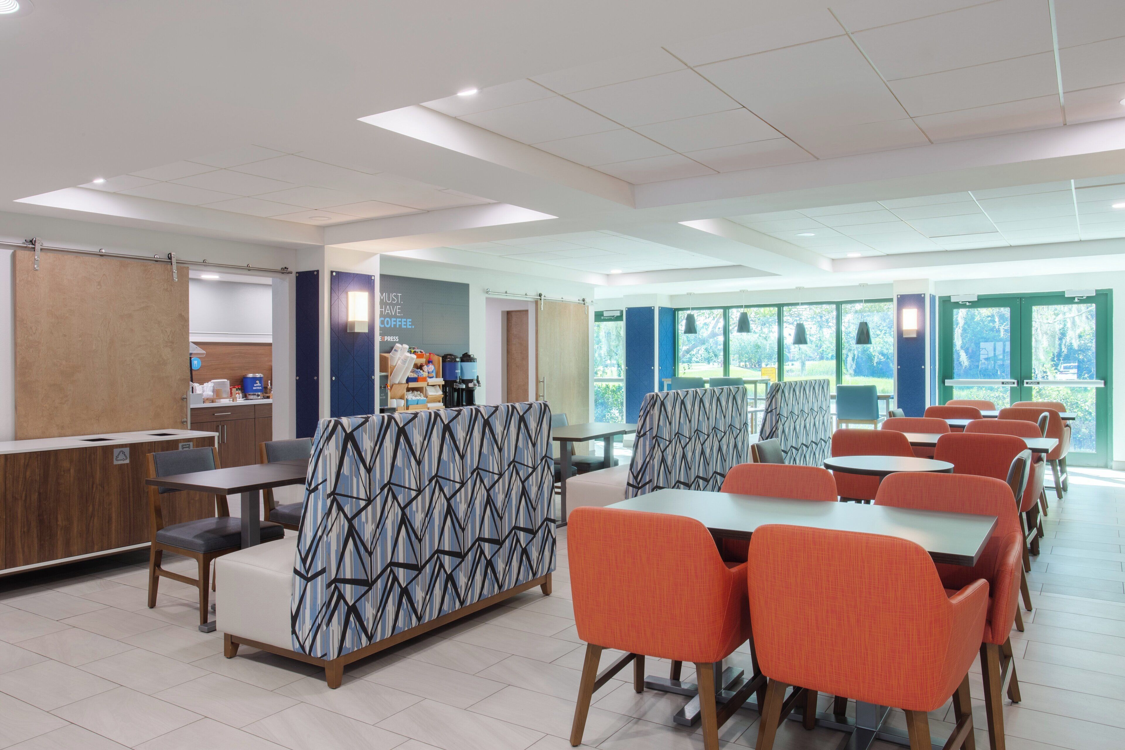 A modern cafeteria with high tables and orange chairs, a counter with food items, and large windows offering a view of greenery.