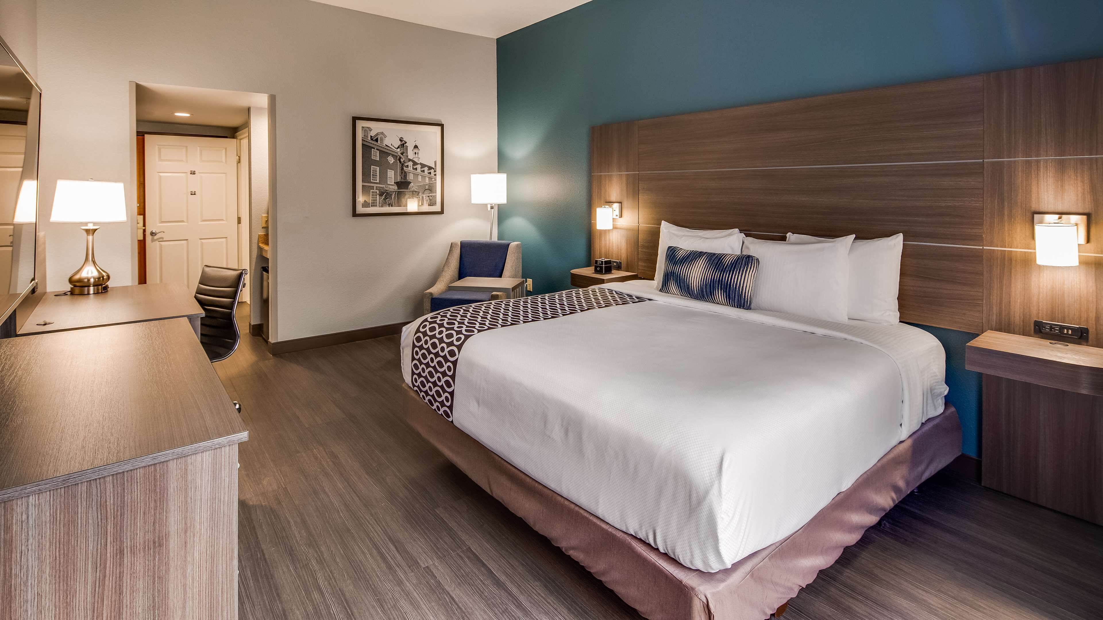 Habitación de hotel con una cama grande, mesitas de noche, un escritorio con lámpara, una silla, un cuadro en la pared y una puerta que da a otra habitación.