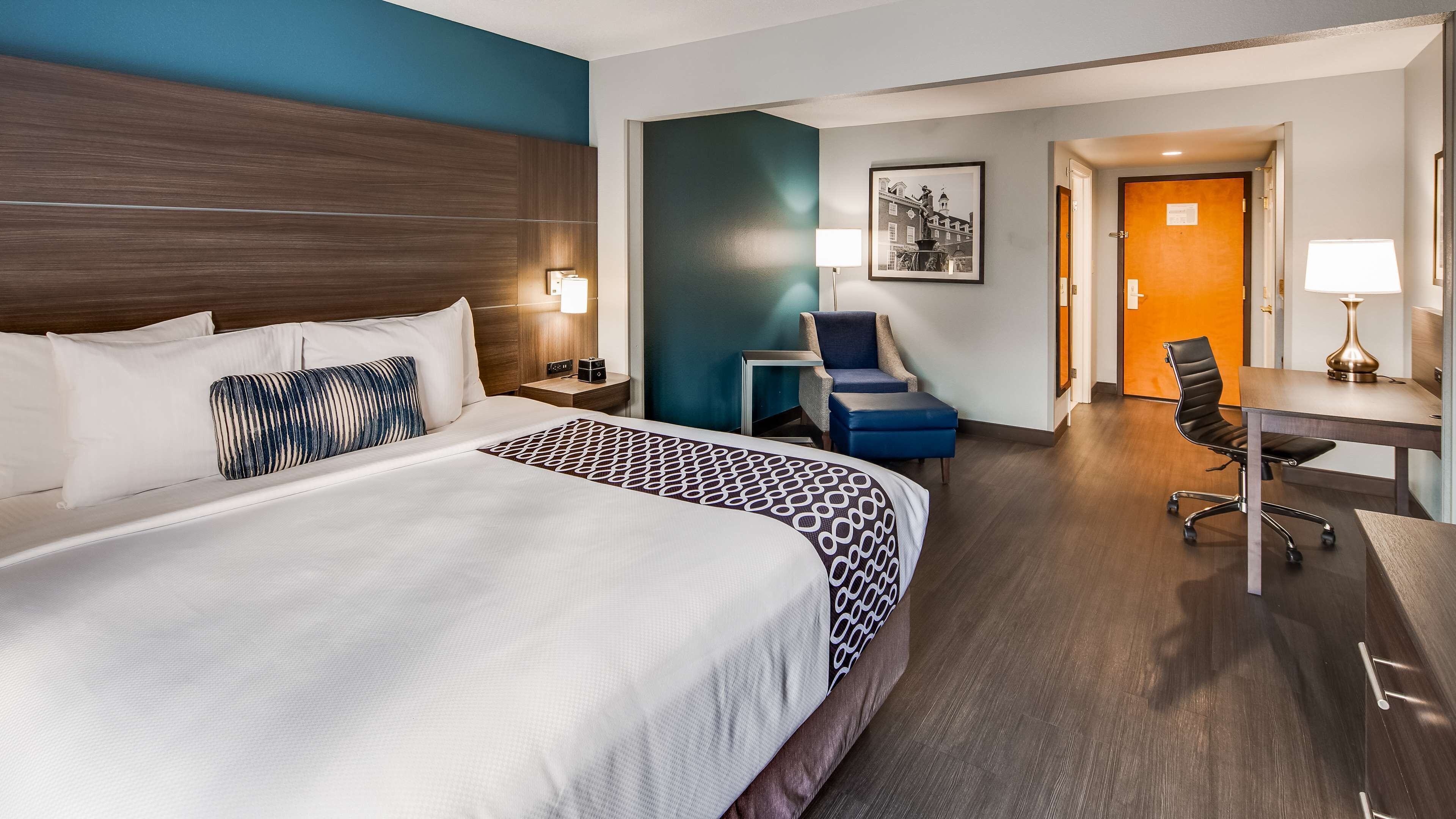 Habitación de hotel con una cama grande, un escritorio, una silla y una mesita de noche con lámpara.