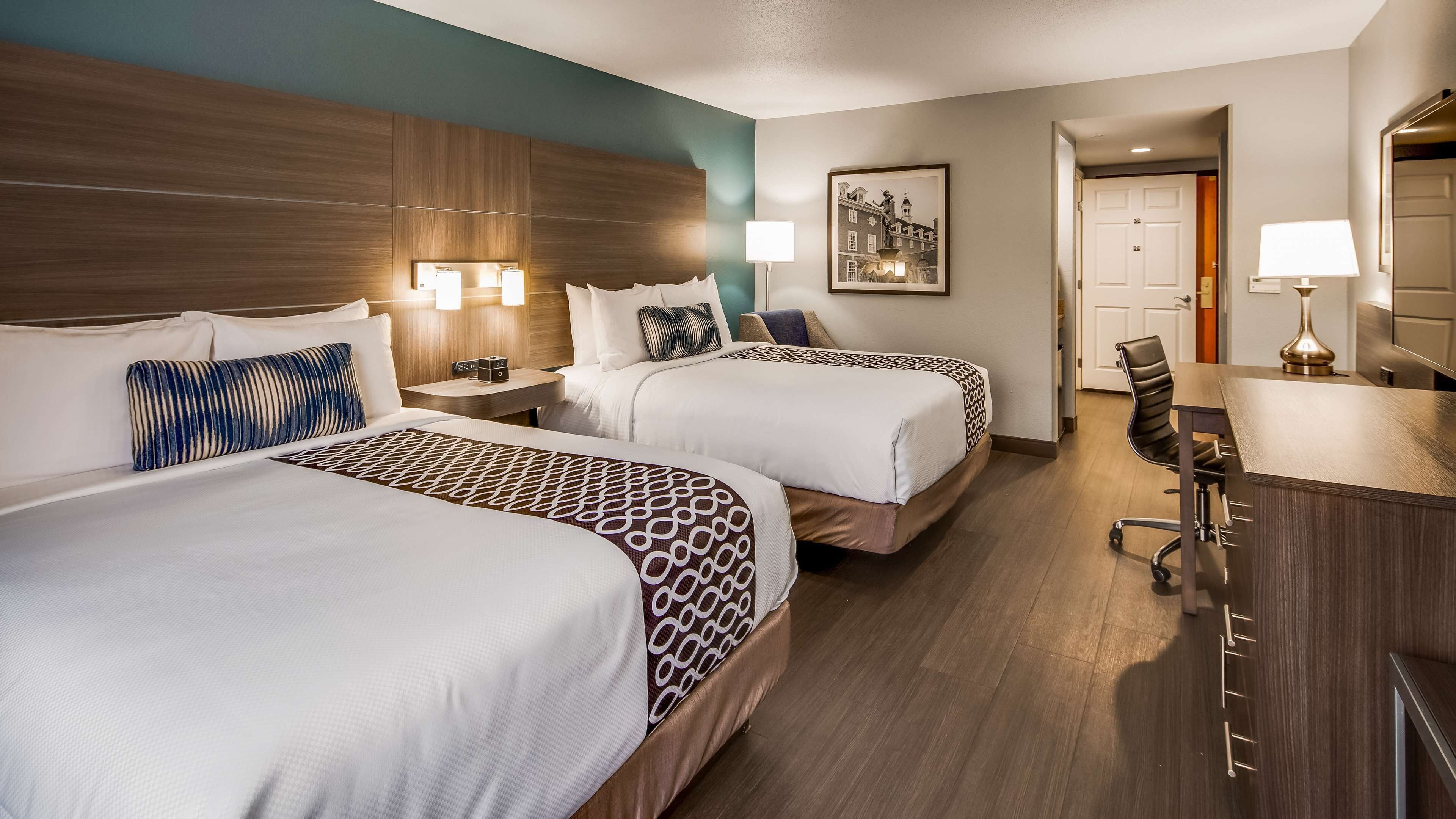Quarto de hotel com duas camas, uma escrivaninha, uma cadeira, um abajur e um quadro na parede.