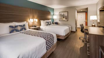 Quarto de hotel com duas camas, uma escrivaninha, uma cadeira, um abajur e um quadro na parede.