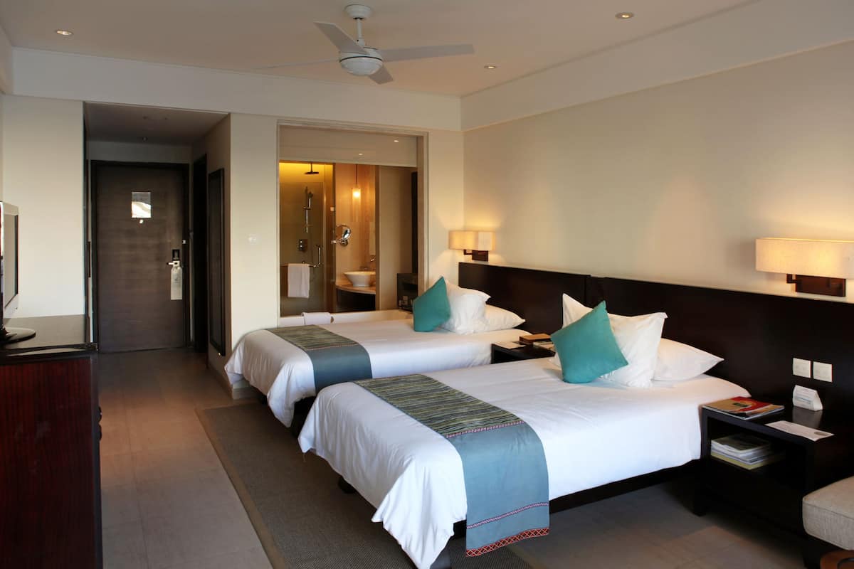 Une chambre d’hôtel avec deux lits, un bureau, une chaise, une télévision et une salle de bain visible à travers une porte ouverte.