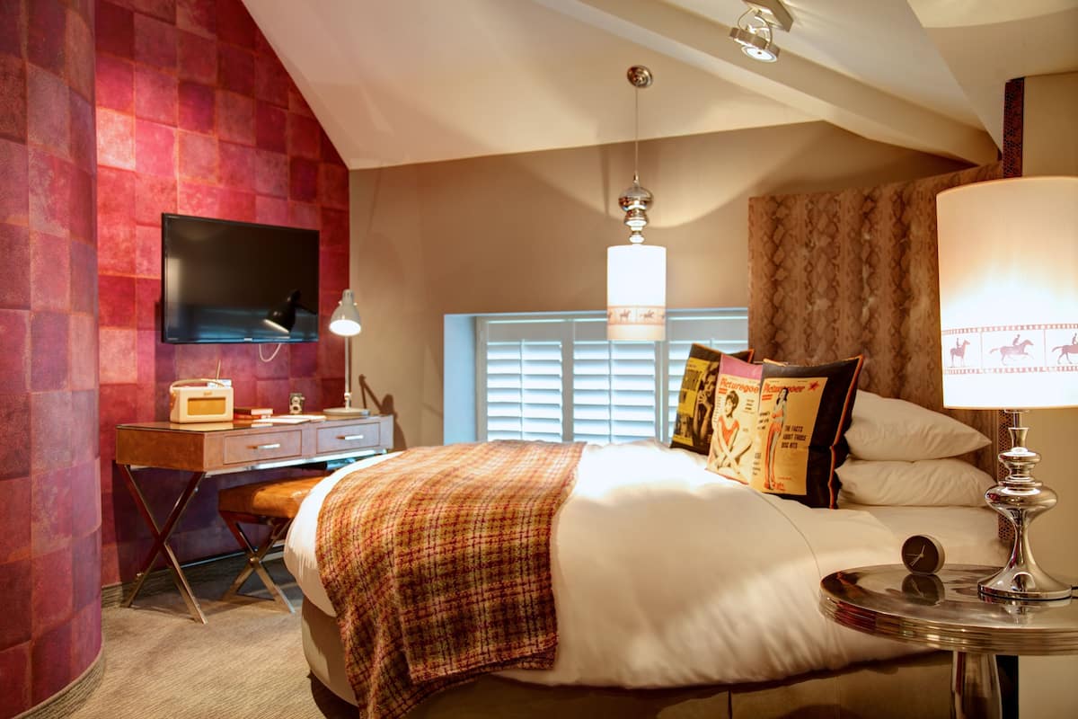 A hotel room with a large bed, a desk with a flat-screen TV, a lamp, and a window with blinds.