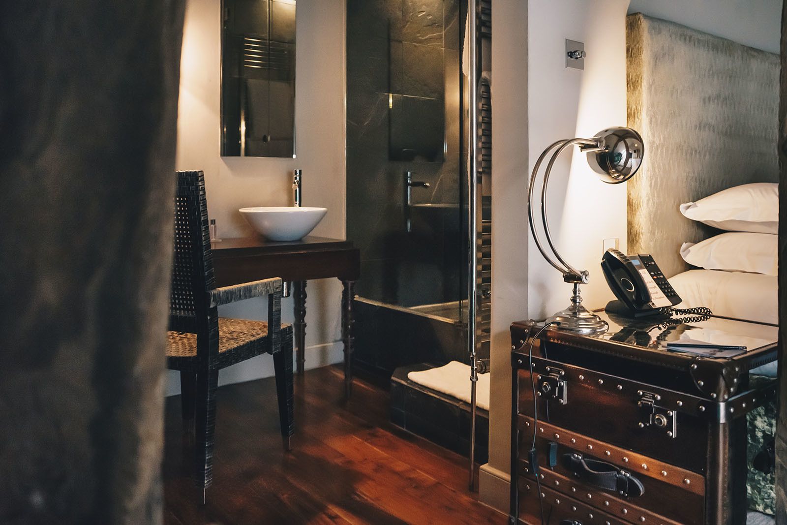 A bathroom with a wooden vanity, a white sink, a mirror, a towel rack, and a suitcase.