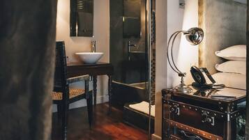 A bathroom with a wooden vanity, a white sink, a mirror, a towel rack, and a suitcase.