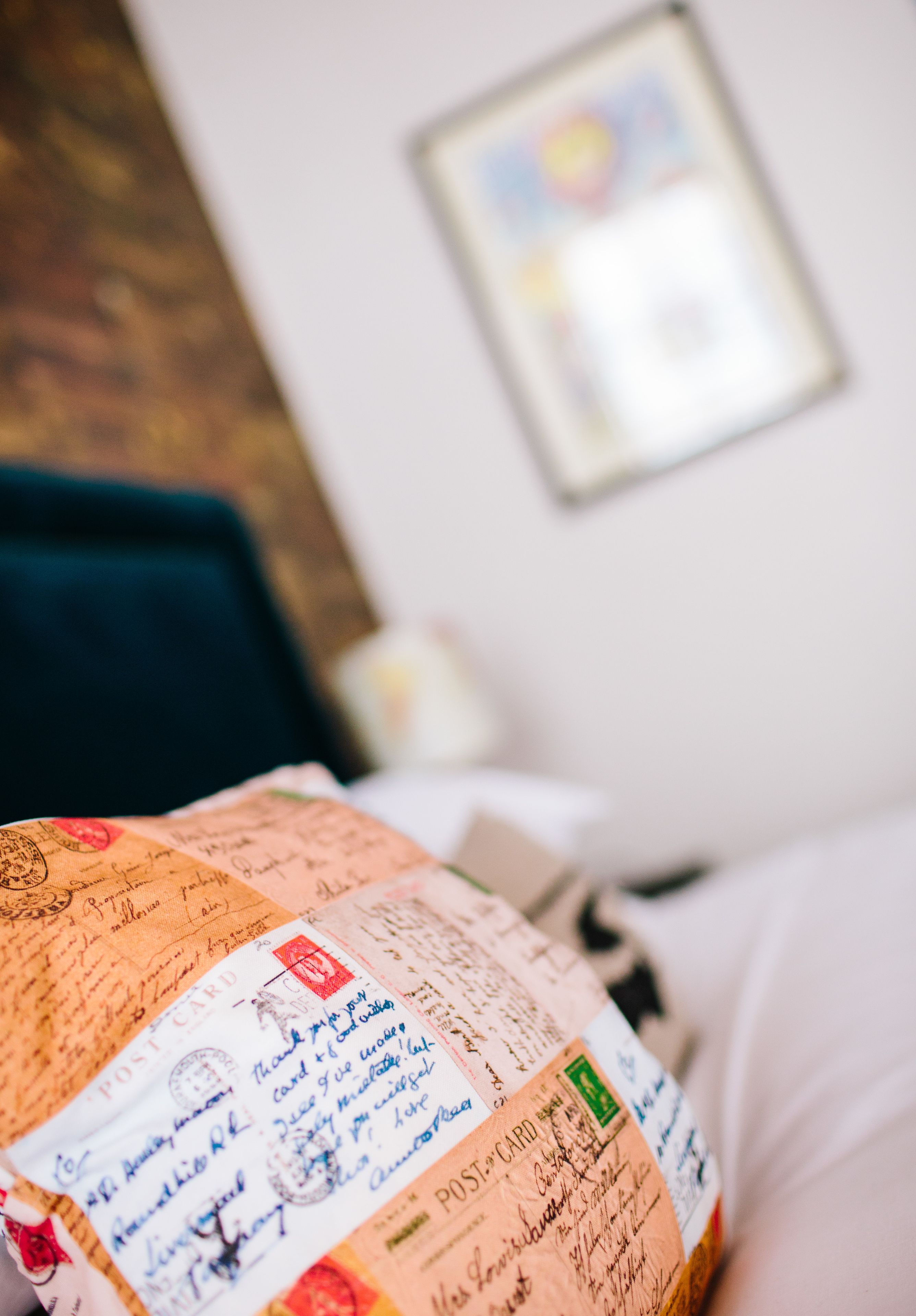A bed with a white pillow and a blanket with a postal card design.