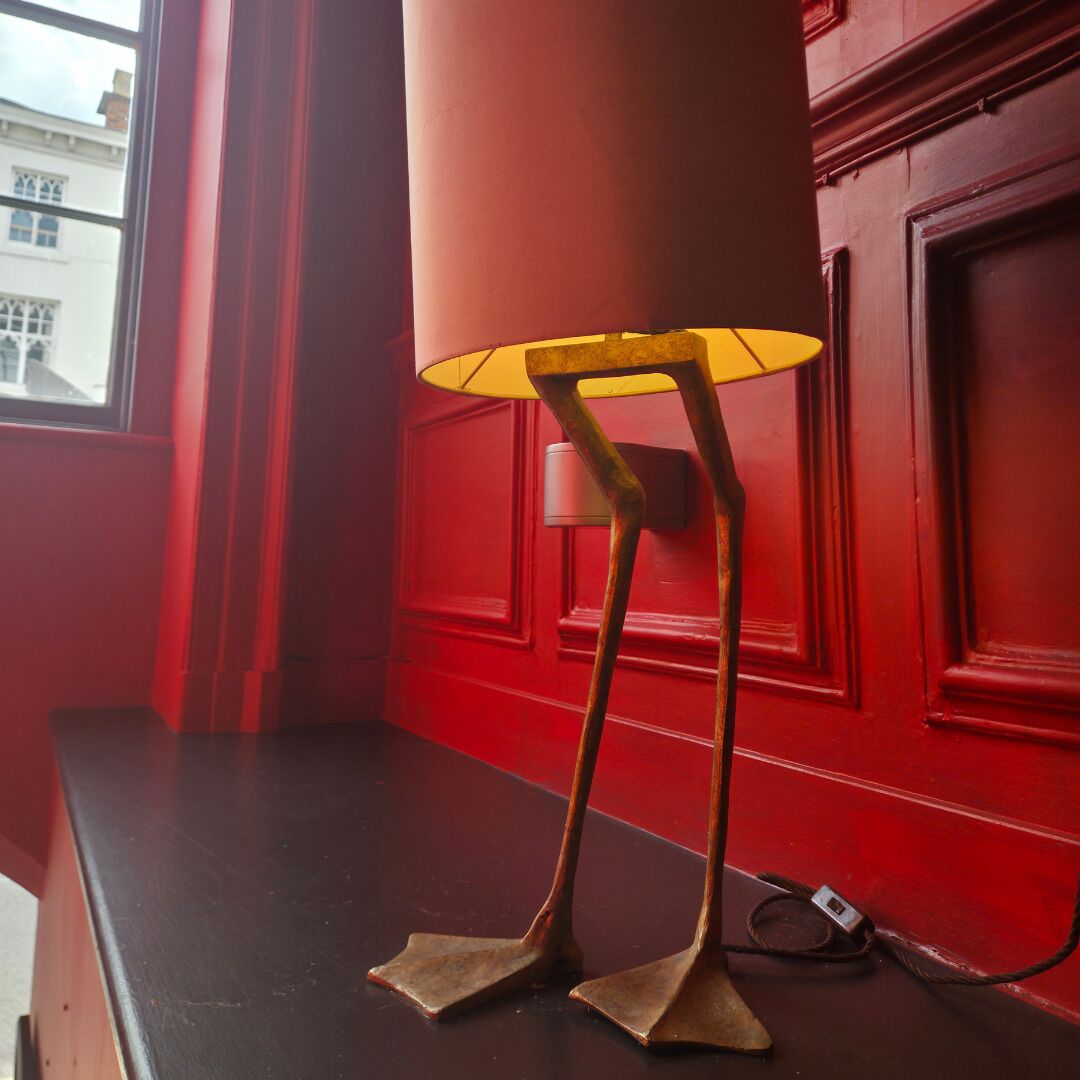 A lamp with a yellow shade on a black surface, next to a red wall with a window.