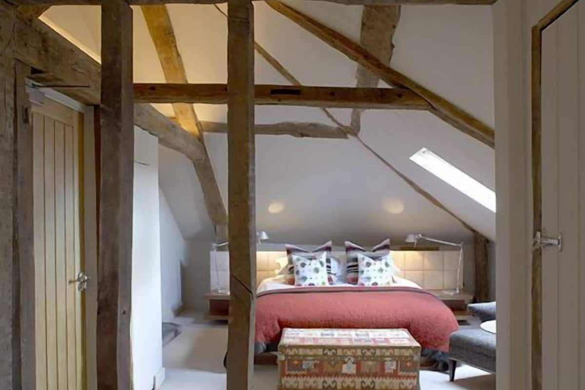 A bedroom with a sloped ceiling, exposed wooden beams, a bed with patterned bedding, a bedside table, and a small chest of drawers.