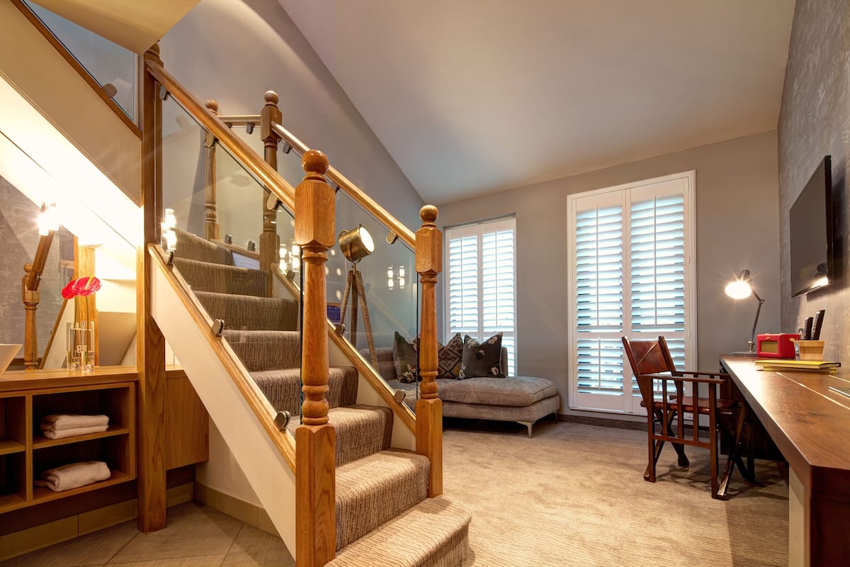 A cozy living room with a wooden staircase, a comfortable sofa, a dining area, and a large window with blinds.