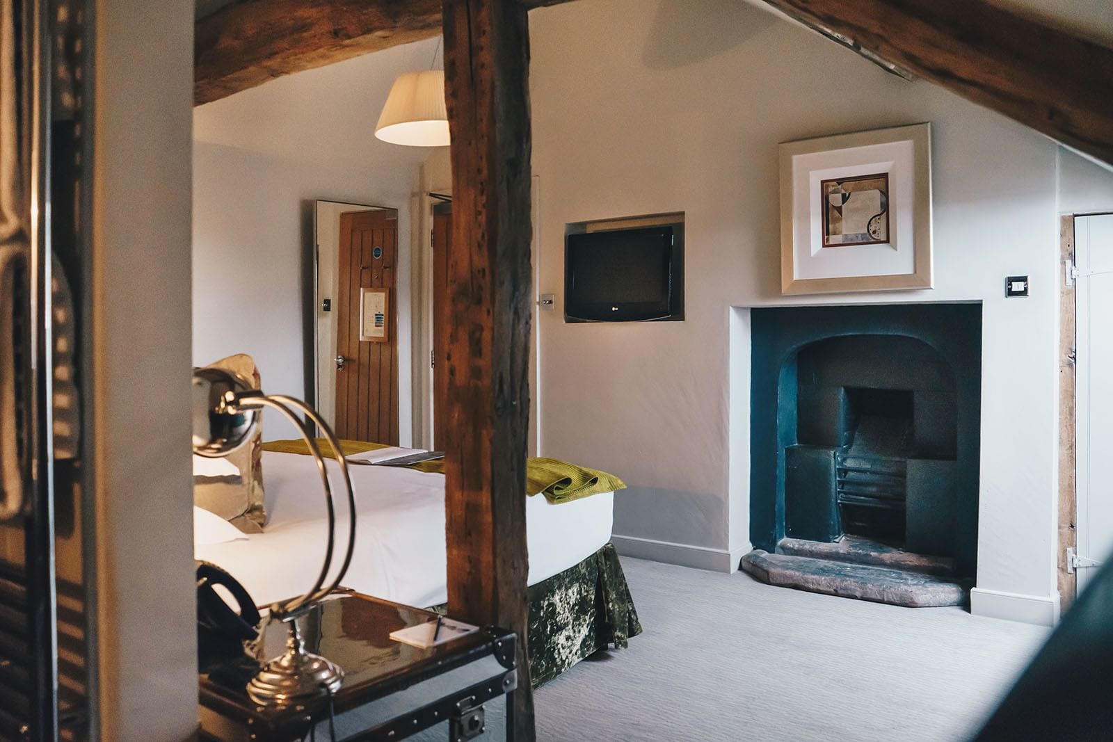 Una habitación con un espejo de madera, un escritorio con una lámpara, una cama con una colcha verde, una chimenea y un televisor en la pared.