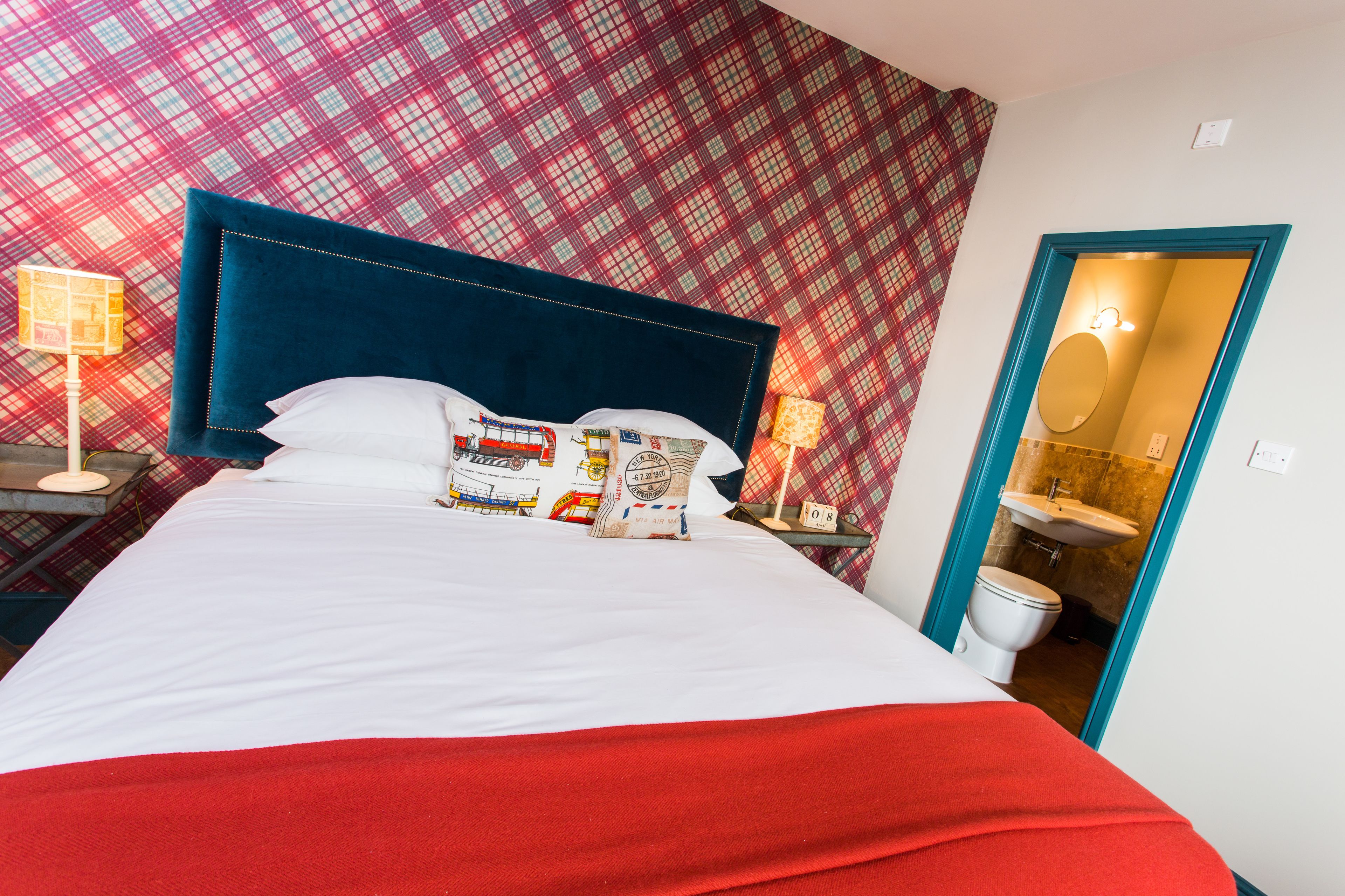 A hotel room with a large bed, a red throw blanket, a headboard with a clock, and a bathroom visible through an open door.
