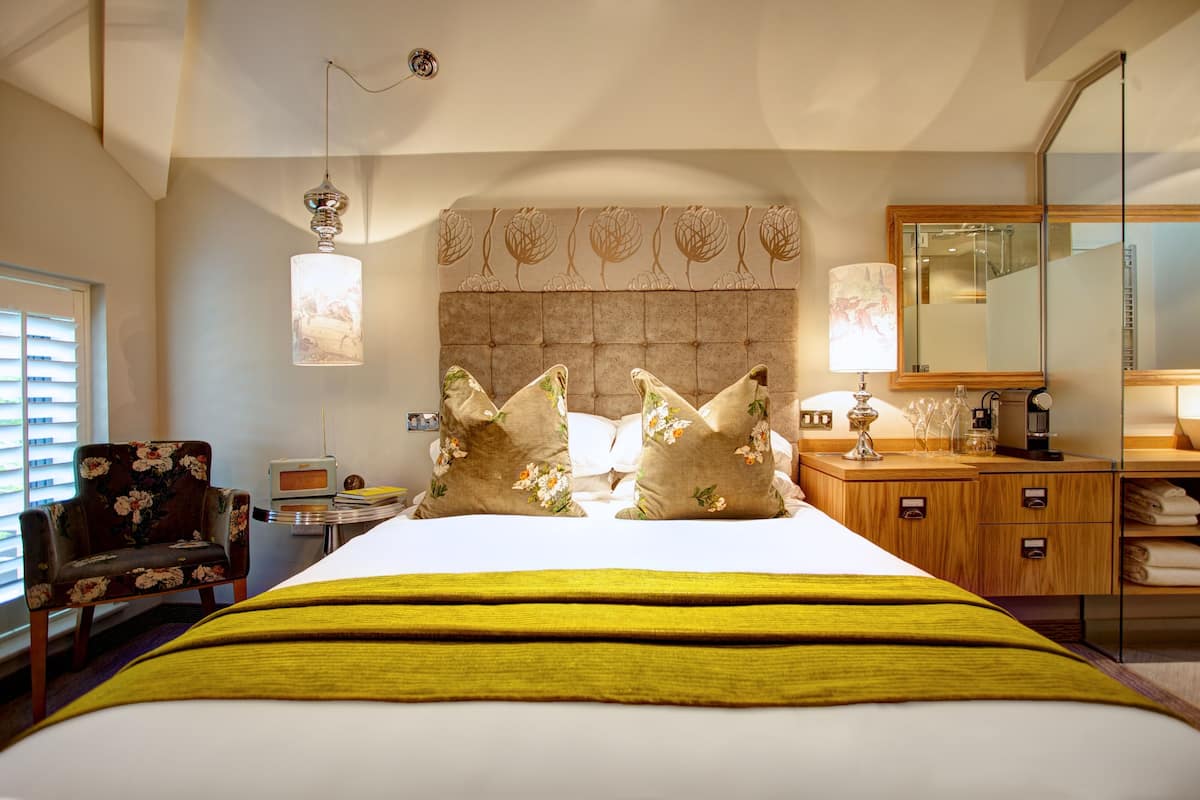 A hotel room with a large bed, a wooden nightstand, a chair, a mirror, and a window with blinds.