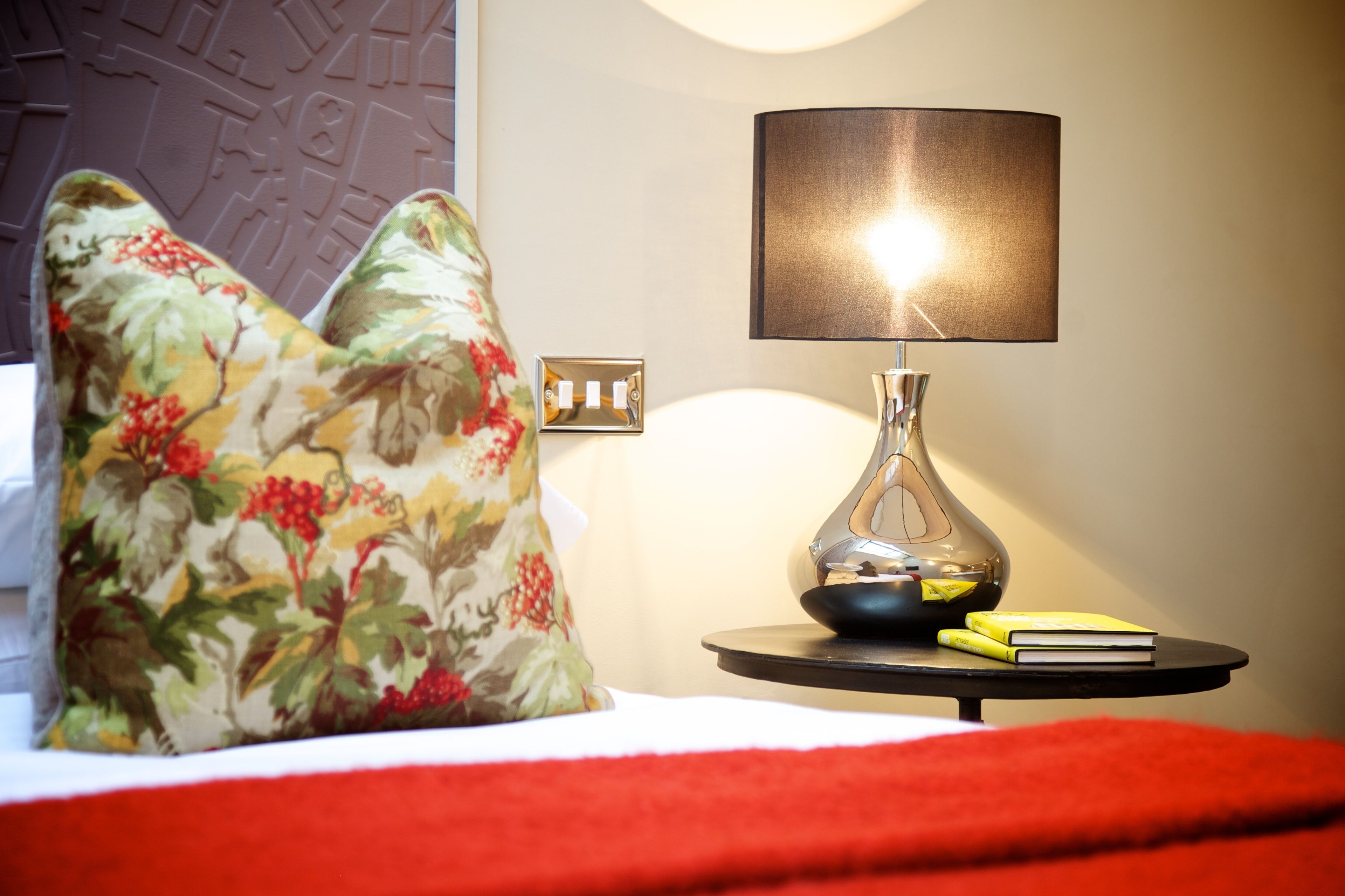 En seng med blomstrede puder, et natbord med en lampe og en bog.