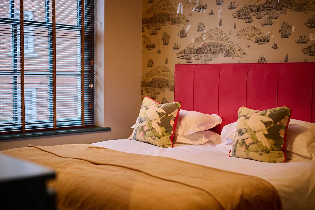 A hotel room with a bed, a window with blinds, and a wall with maritime-themed wallpaper.