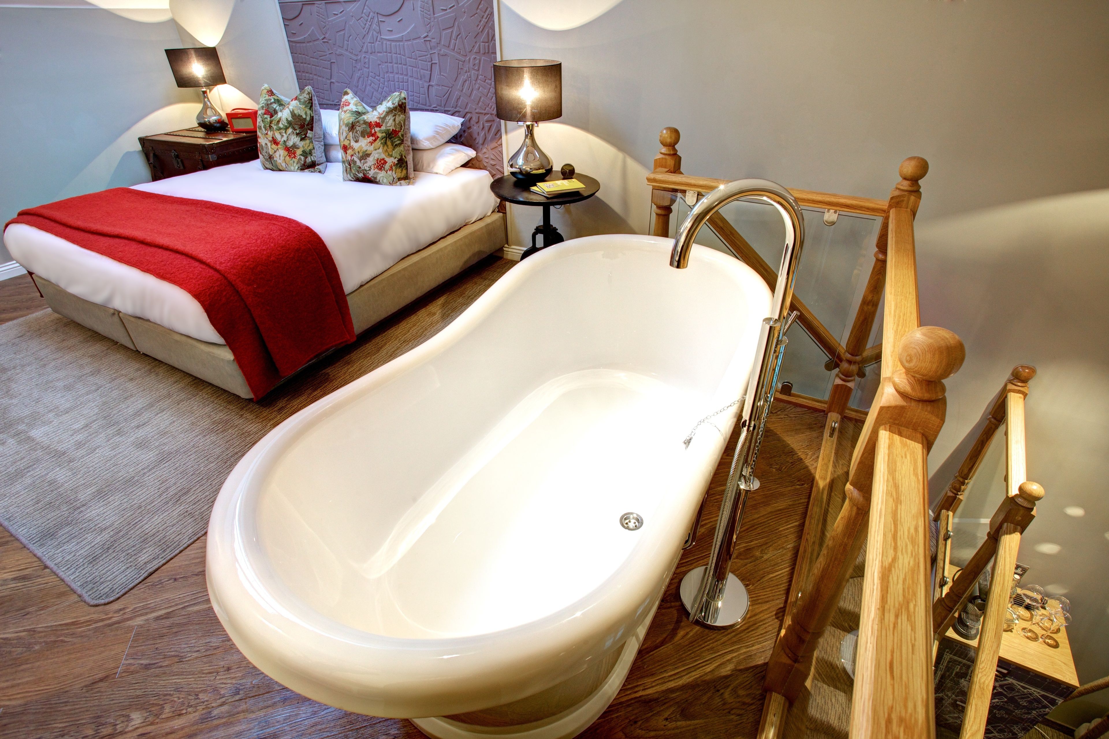 A bedroom with a large bathtub, a bed with a red blanket, two bedside tables with lamps, and a wooden staircase.