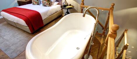 A bedroom with a large bathtub, a bed with a red blanket, two bedside tables with lamps, and a wooden staircase.