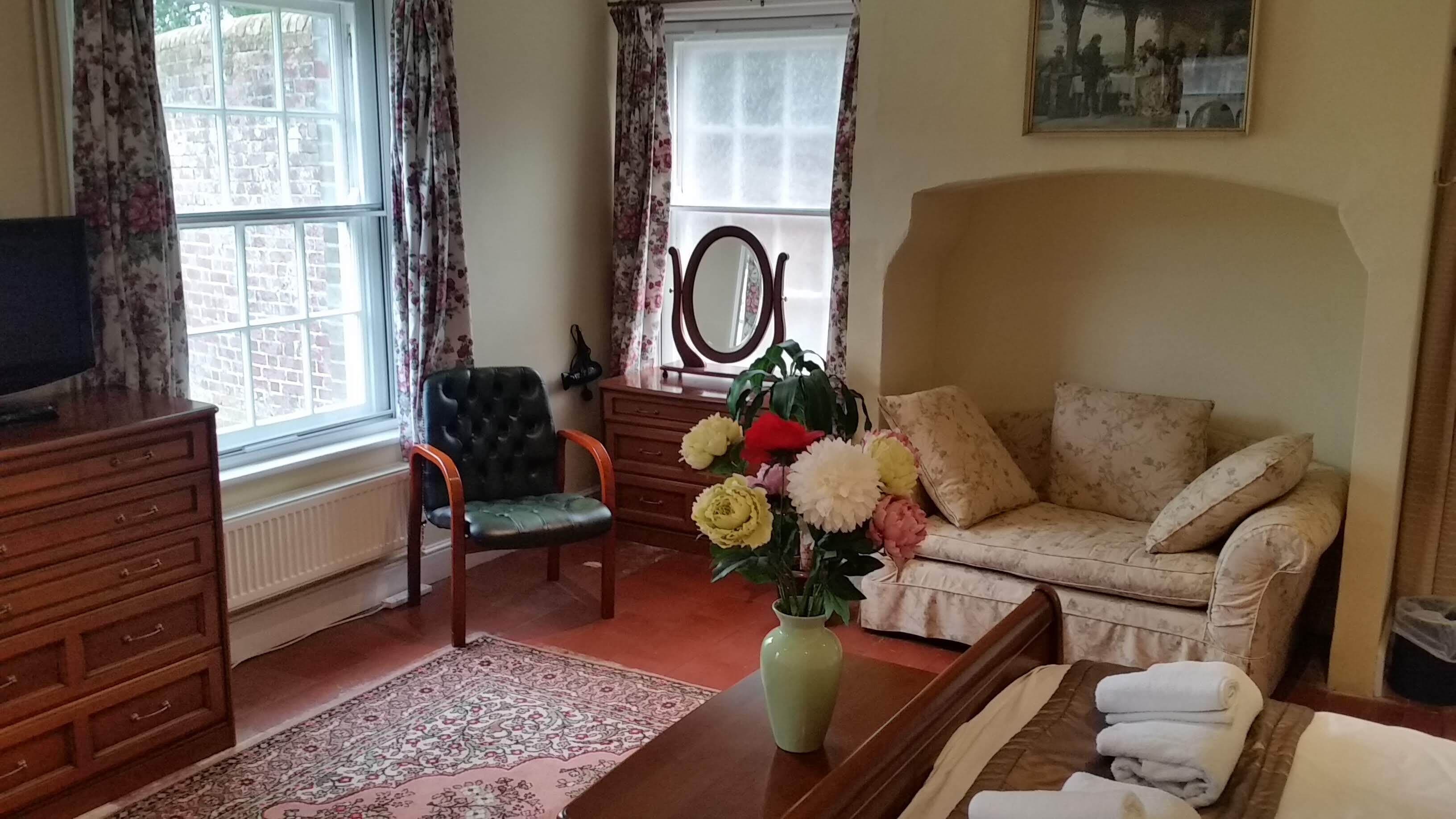 Una habitación con sofá, sillón, cómoda, televisor, espejo, un jarrón con flores y ventana con cortinas.