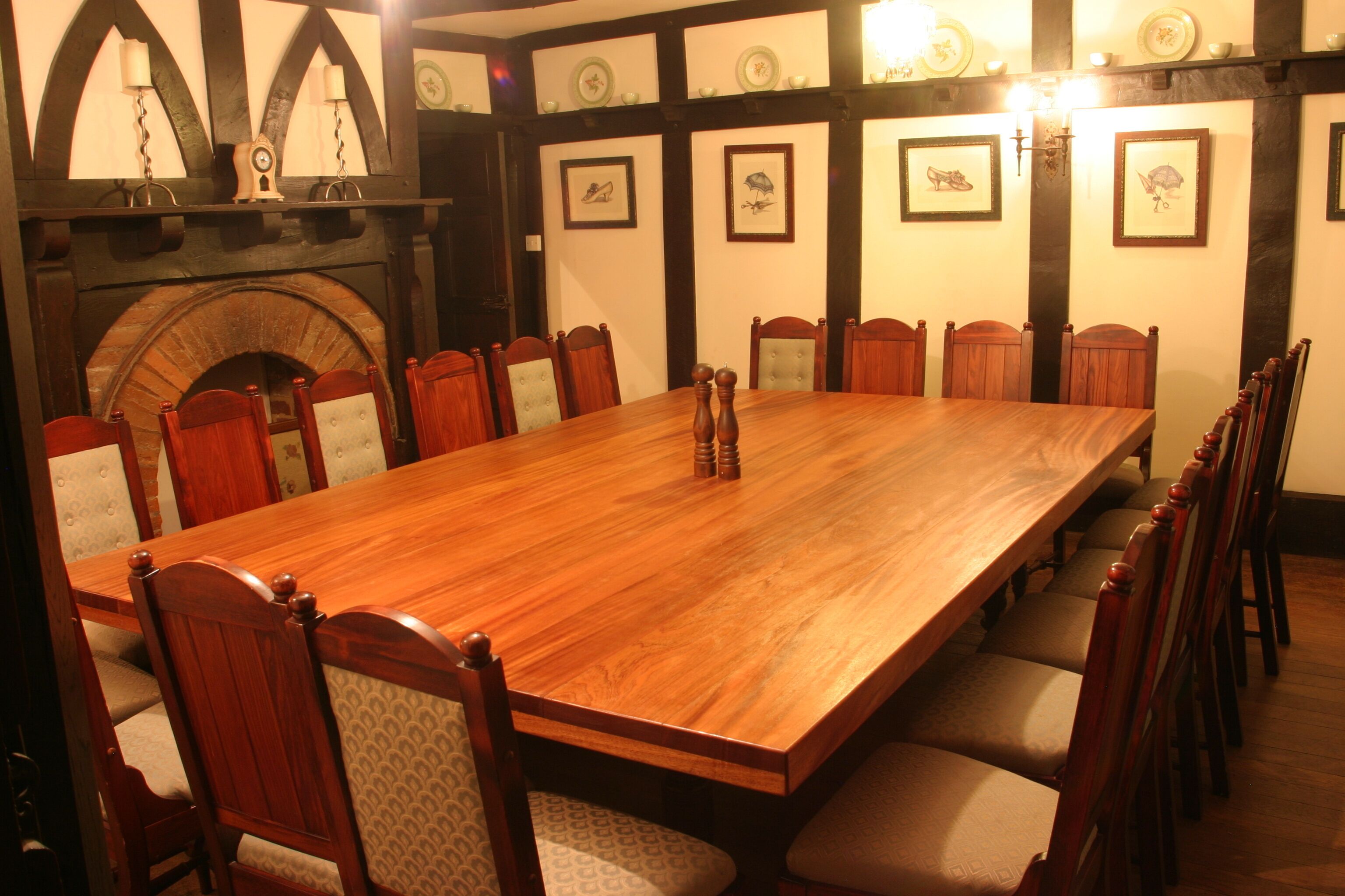 A long wooden dining table surrounded by wooden chairs with cushioned seats, set in a room with a fireplace and framed pictures on the walls.