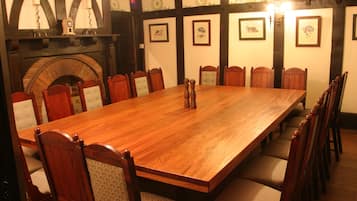 A long wooden dining table surrounded by wooden chairs with cushioned seats, set in a room with a fireplace and framed pictures on the walls.