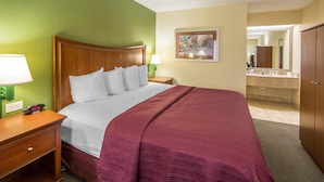 Una habitación de hotel con una cama grande, una mesita de noche con lámpara, un cuadro en la pared y un baño visible a través de una puerta abierta.