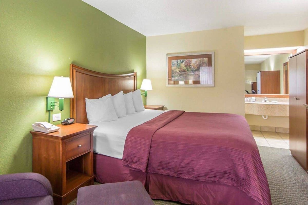 A hotel room with a bed, bedside lamps, a nightstand, a desk, a chair, and a bathroom visible in the background.