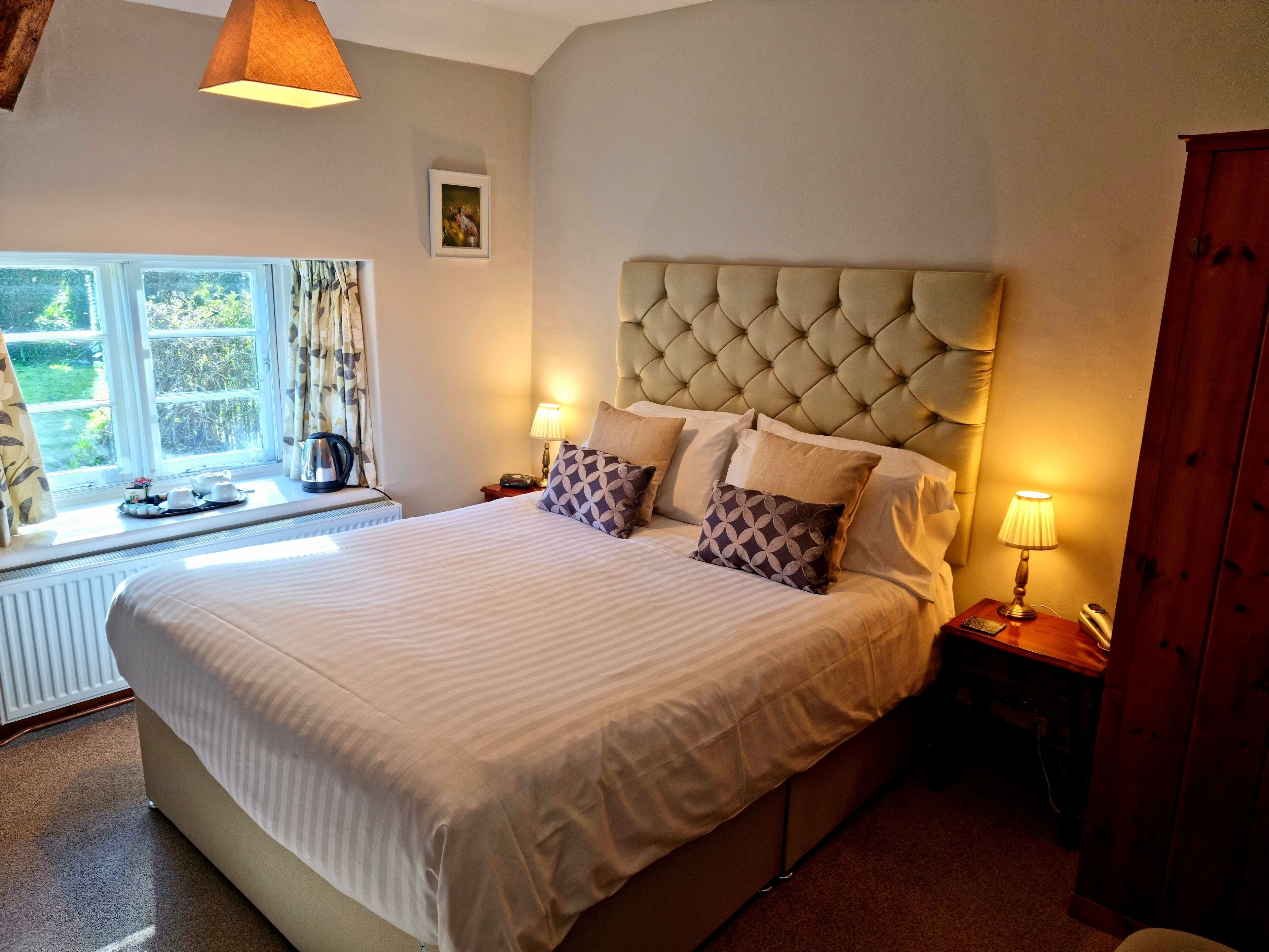 A well-lit bedroom with a large bed, a window with curtains, a bedside table with a lamp, and a radiator.