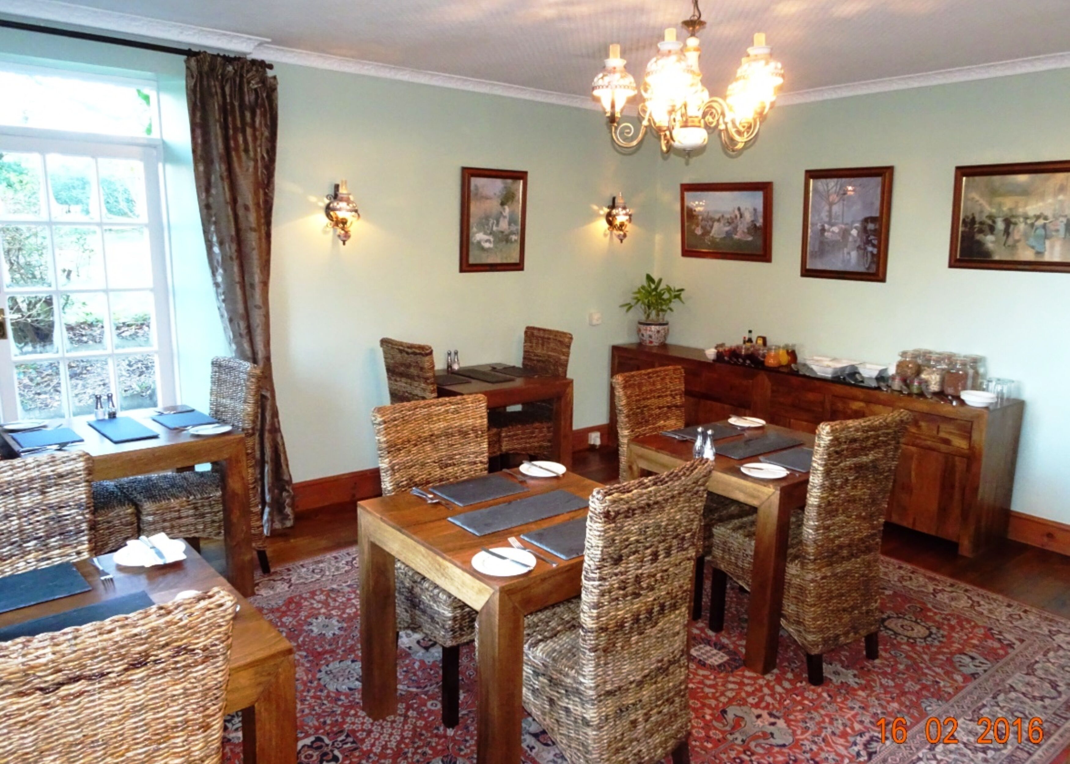 Un'area pranzo con sedie in vimini, tavoli in legno e un lampadario. Alle pareti sono presenti dipinti e una finestra con tende.