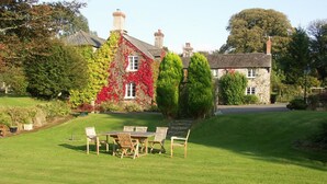 A well-maintained lawn with a wooden table and chairs in the foreground, surrounded by stone buildings with ivy and red flowers. - Collaven Manor Hotel (Okehampton)